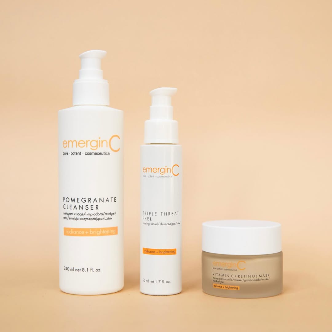 Three emerginC® Radiance + Brightening products. Pomegranate Cleanser, Triple Threat Peel, and Vitamin C + Retinol Mask - displayed on a soft peach background. Clean, minimal composition reflecting clinical skincare and radiant skin results.